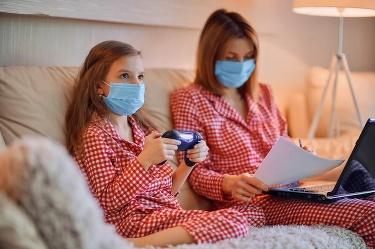 Kobieta w piżamie pracująca z maską, a córka gra w gry komputerowe